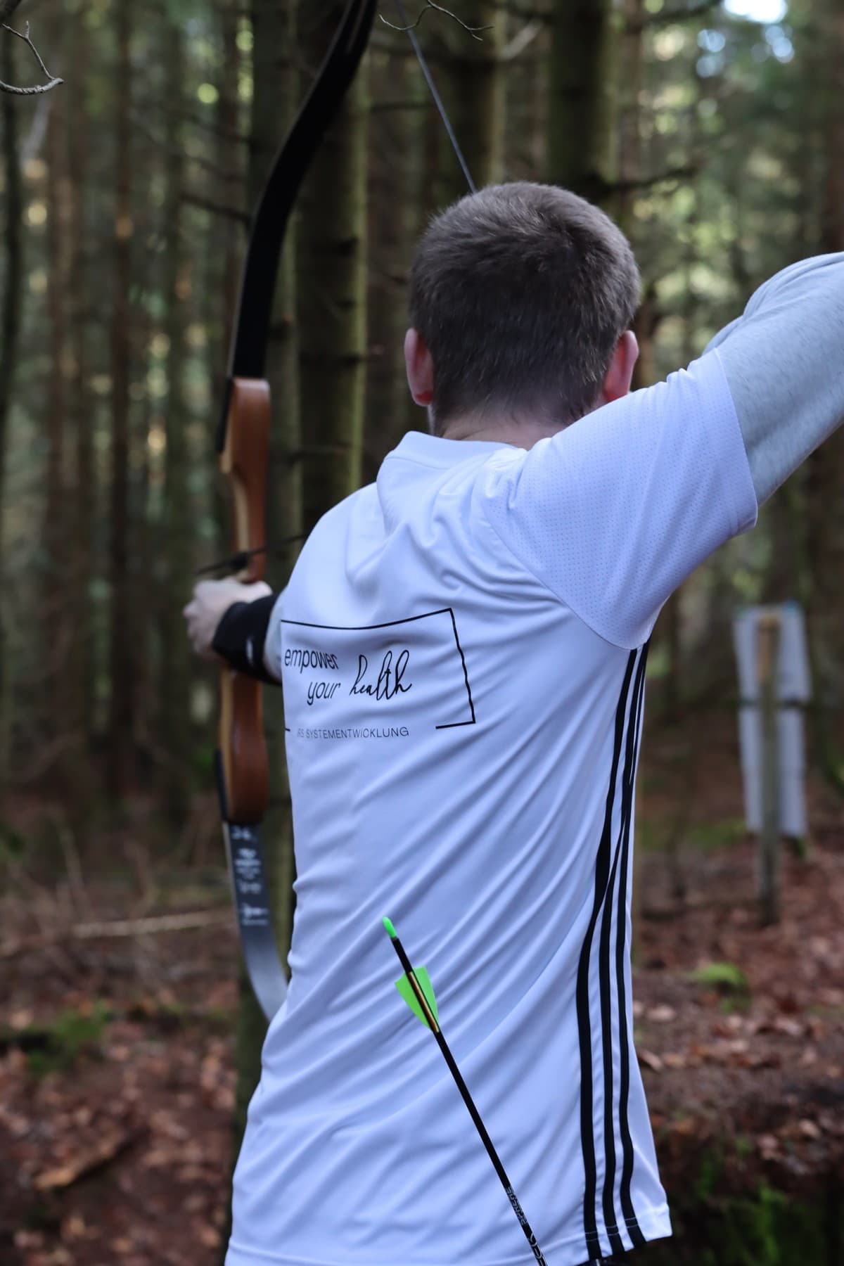 Mitarbeiter beim Bogenschießen im Wald, Shirt mit Empower-your-Health-Aufdruck